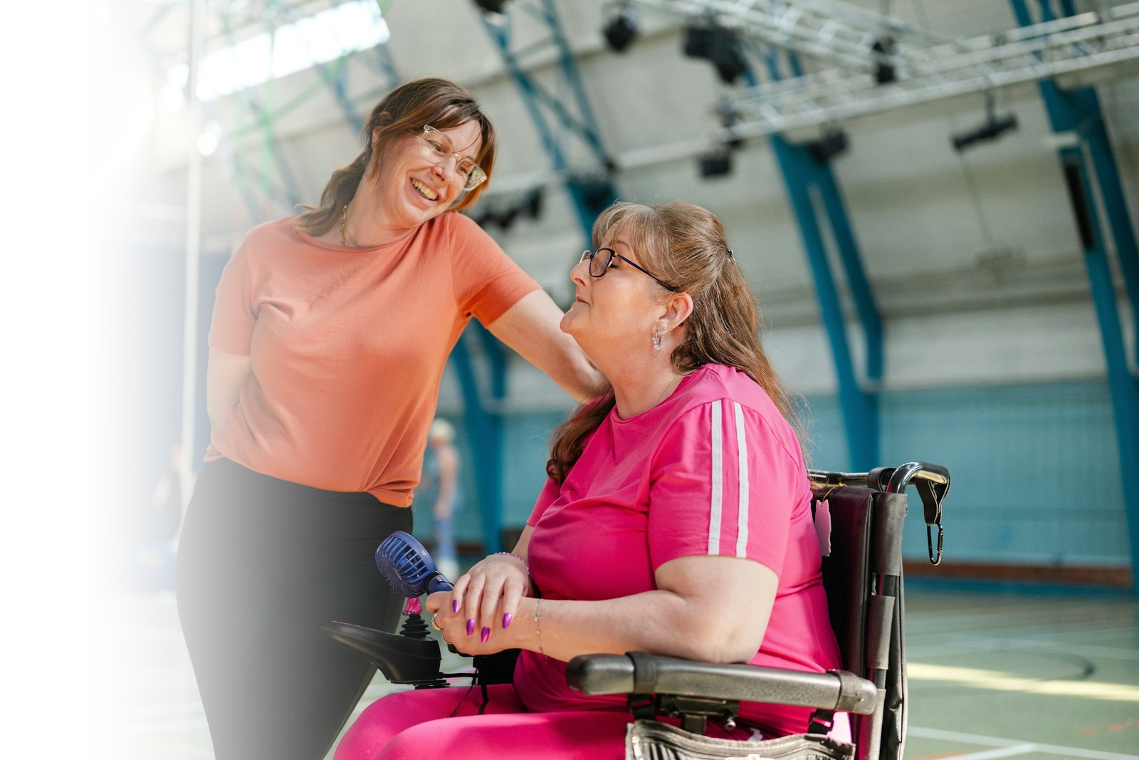 Two women laughing together at a community sports session, one of them using a power wheelchair